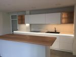 Frente de cocina lineal blanco con iluminación LED técnica continua y encimera de isla en madera natural.