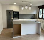 Cocina con isla compacta en blanco y gris, iluminación colgante de diseño y almacenamiento lateral.