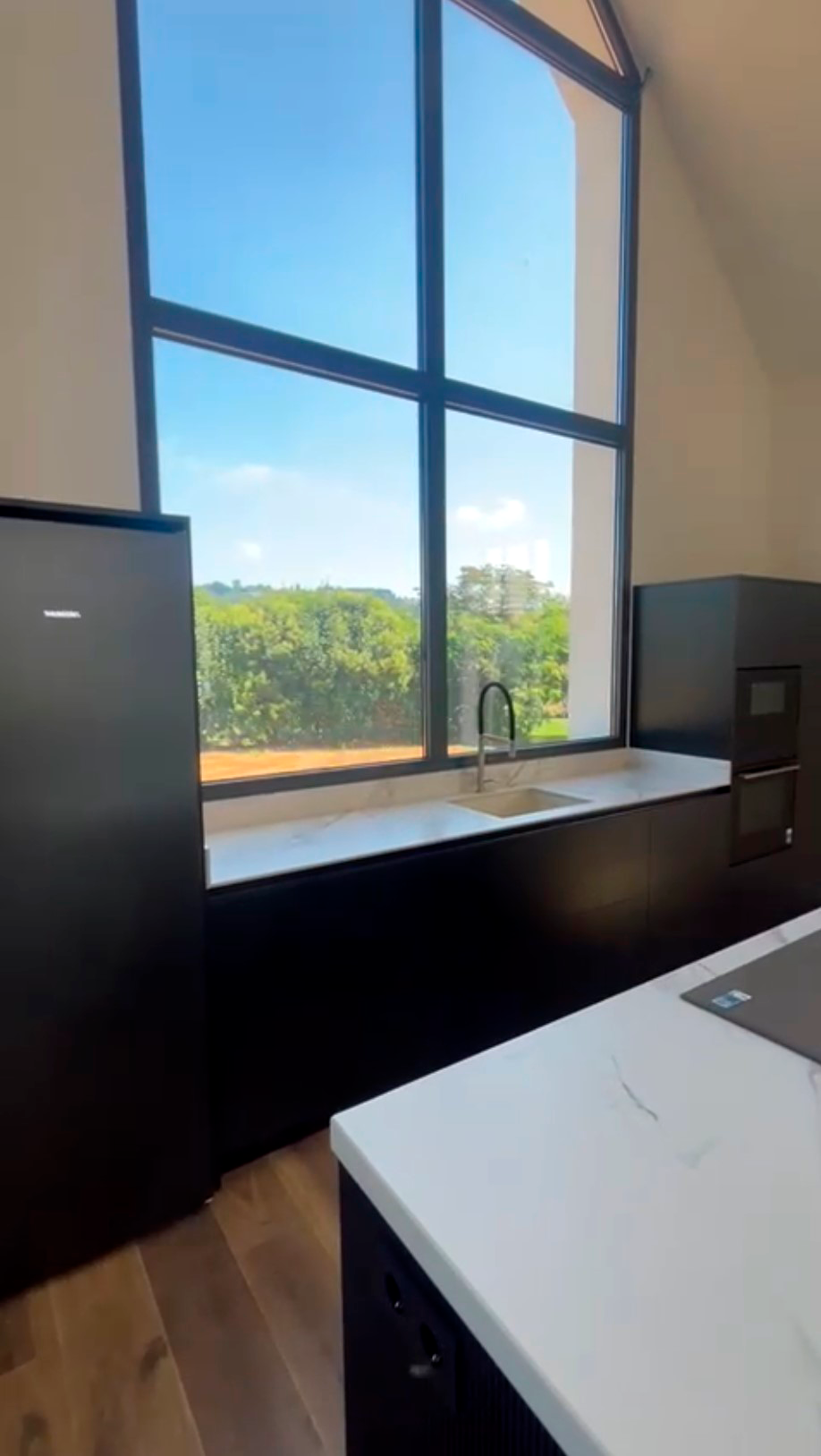 Cocina negra lineal con encimera marmoleada y fregadero bajo un ventanal panorámico.