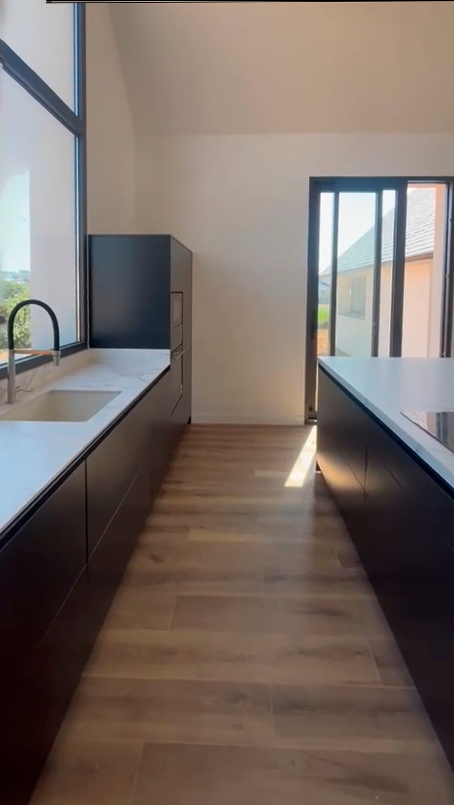 Cocina negra lineal con encimera marmoleada y fregadero bajo un ventanal panorámico.